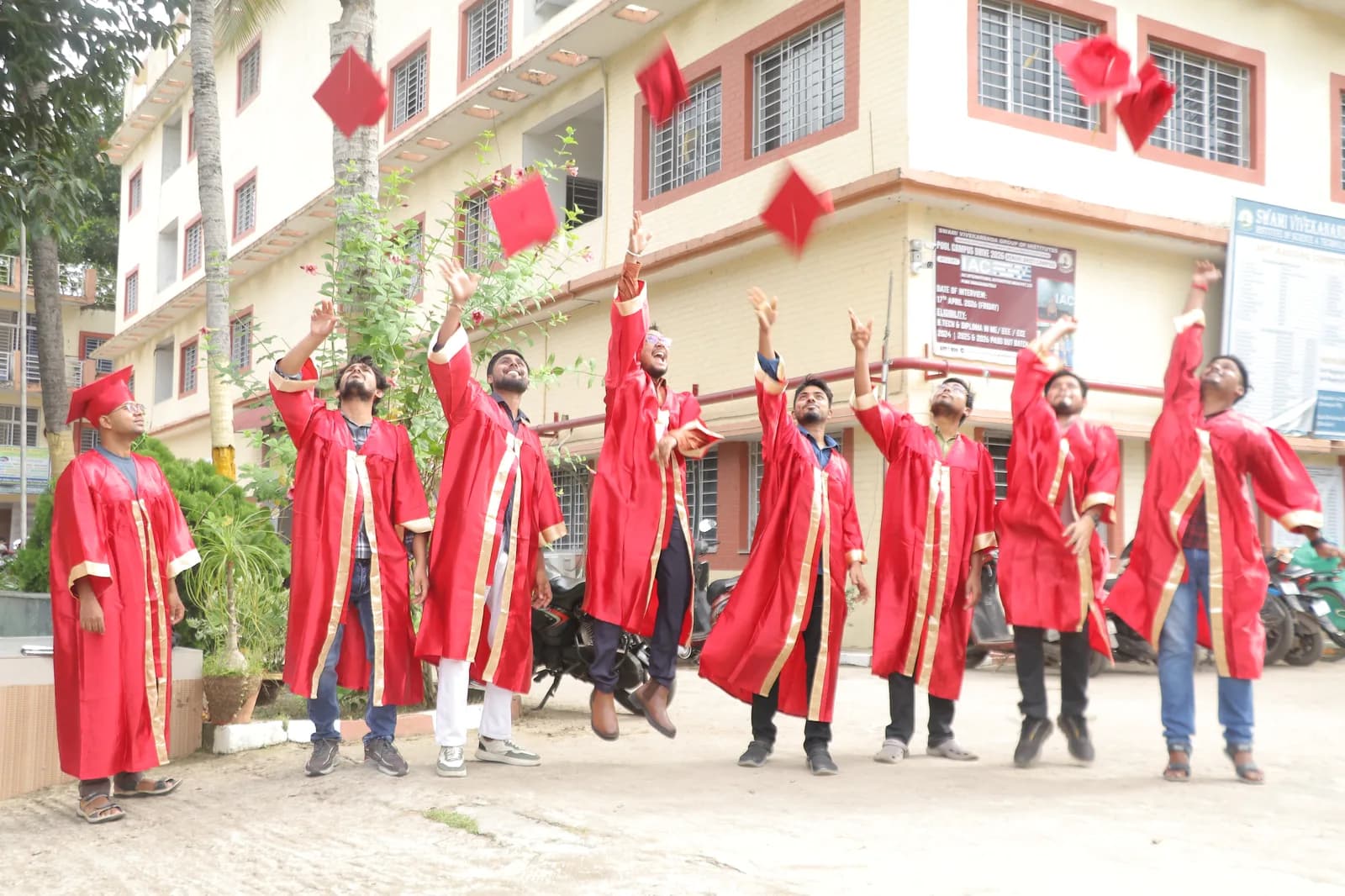 A group of SVIST B.Tech graduates in red and gold graduation gowns throwing their caps into the air in celebration outside the campus building during Graduation Day