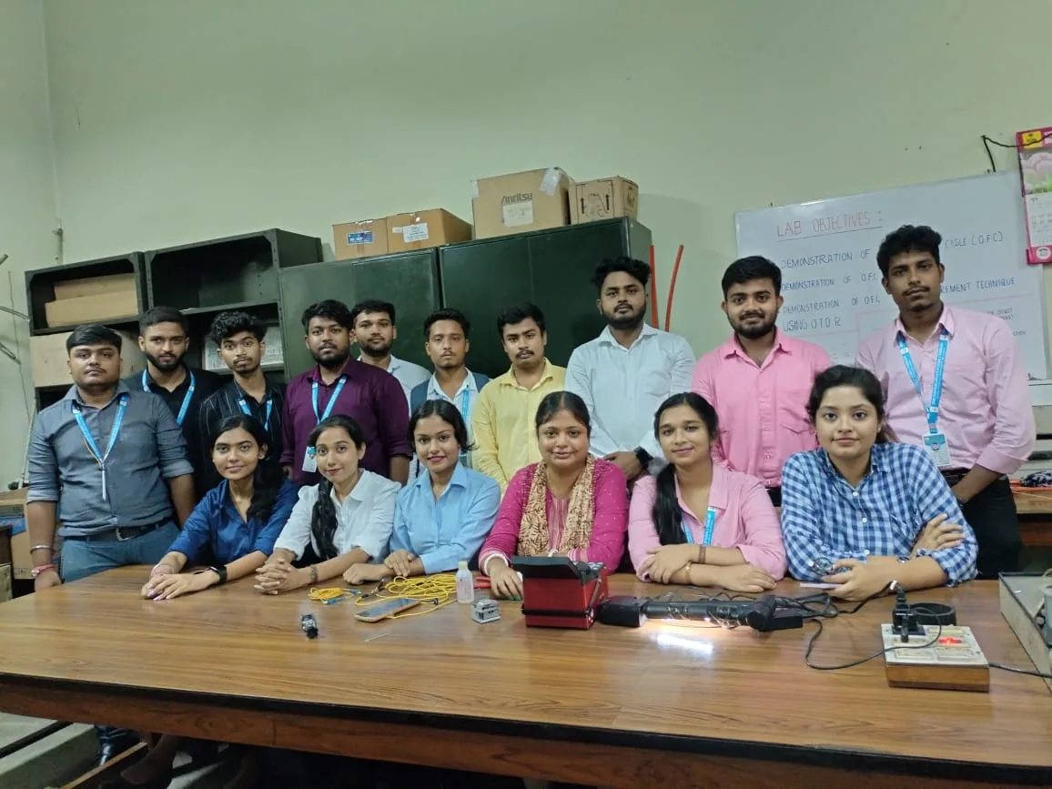 SVIST students and faculty seated around a table with optical fibre splicing equipment during an industrial visit to a BSNL lab, with a whiteboard listing OFC demonstration objectives in the background