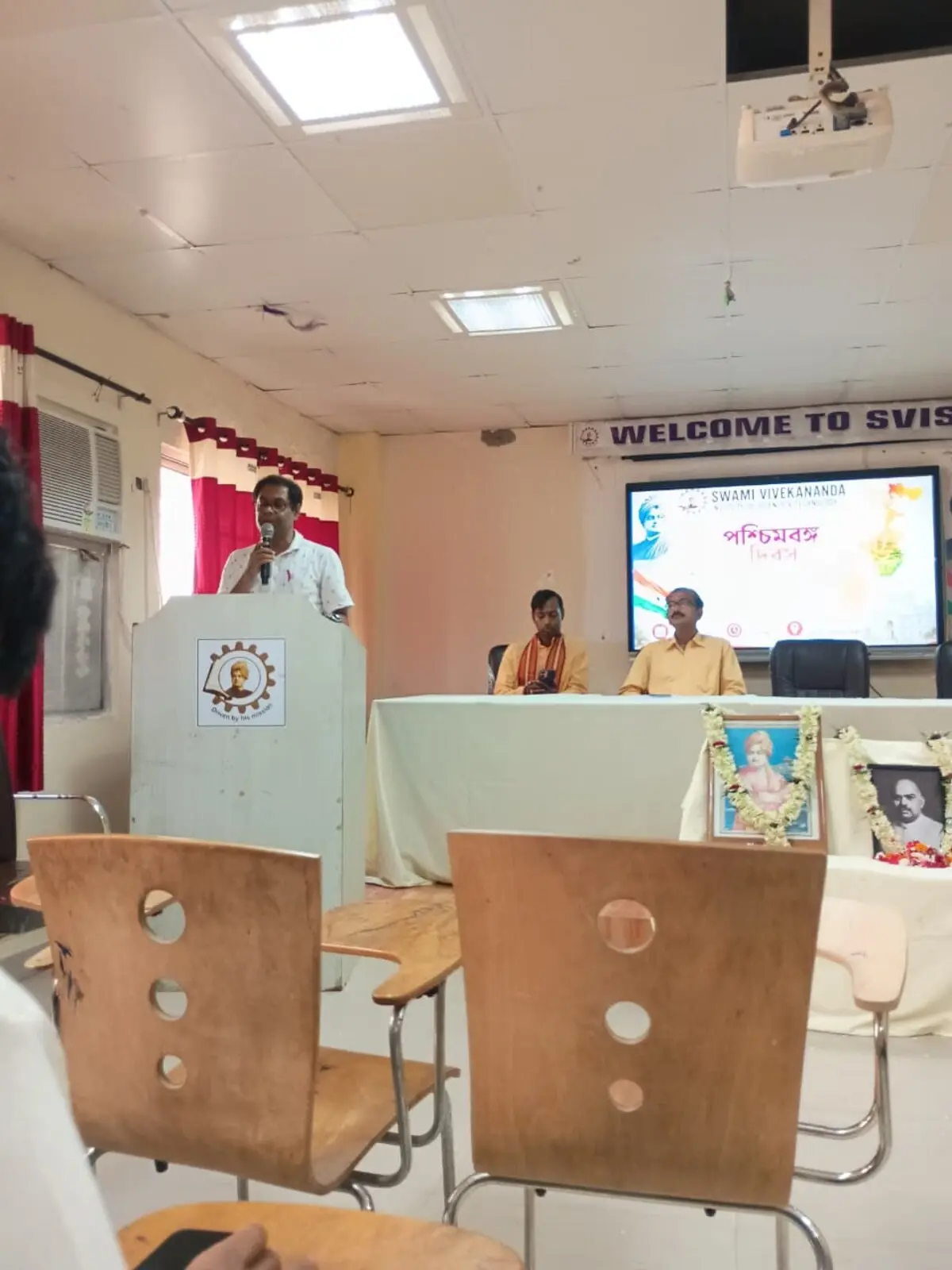 Faculty member addressing the audience at the podium during the Paschim Banga Dibas seminar at SVIST, with a screen behind showing the Paschim Banga Dibas title slide